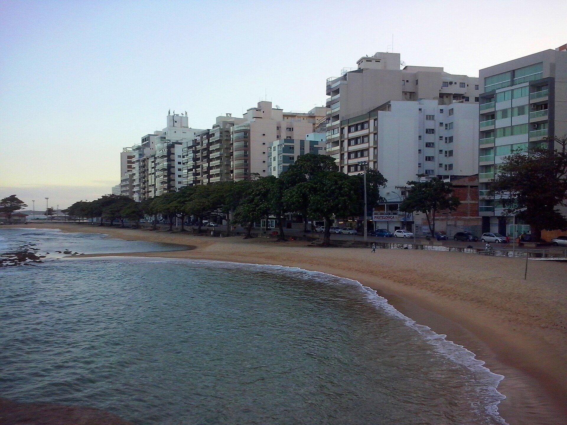 Vista da Praia da Castanheira no Centro de Guarapari, com árvores na orla, pedras e água calma.