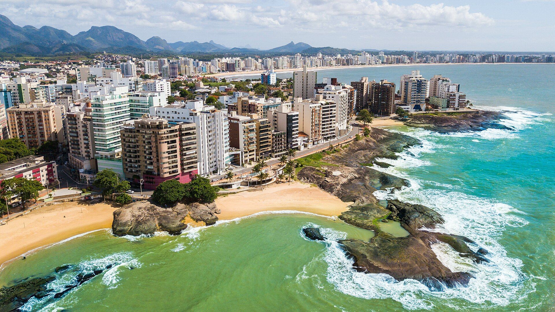 Vista aérea de Guarapari mostrando a faixa costeira, os bairros urbanos e o relevo ao fundo.